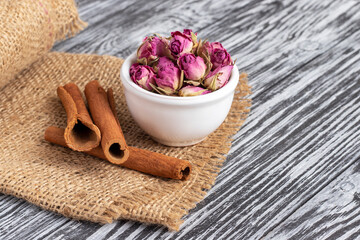Dried damask rose buds in small white bowl and cinnamon sticks on black wooden table.