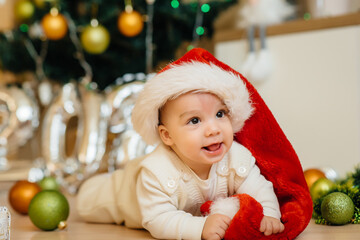 Cute smiling baby is lying under a festive Christmas tree and playing with gifts. Christmas and New Year celebrations
