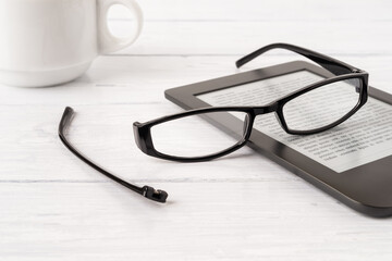 Eyeglasses with one temple broken off the rim on a e-reader over white wood desk. Breakage of...