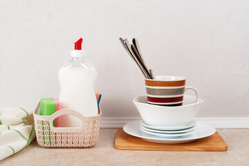 White dishwashing liquid and few foam sponges in a small basket near a stack of clean dishes on the kitchen table against white concrete wall. Household kitchen detergent.