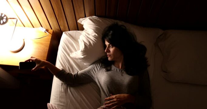 Woman lying in bed turns off smartphone and bedside nightstand lamp