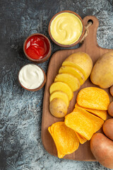 Vertical view of crispy chips and uncooked potatoes on wooden cutting board and mayonnaise and ketchup on gray table