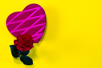On a yellow background, there is a beautiful purple heart with crossed pink fabrics. Next to it, a beautiful red rose tucked into a small glass bottle. Copy space to the right of the image