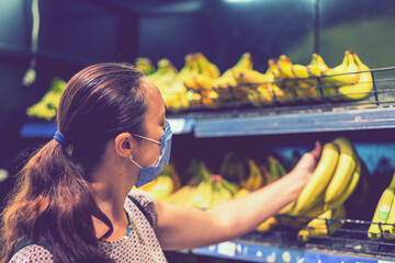 Focused woman in face mask choosing fruits, taking bananas from shelves in grocery store. Customer in supermarket. Side view. Shopping during epidemic concept. toned