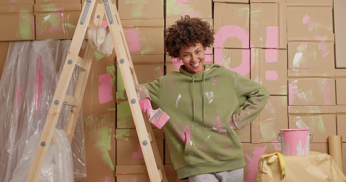 Smiling young Afro American woman engaged in painting walls leans hand on wooden ladder does reconstruction of new dwelling renovates house stands dirty. Relocation home improvement concept.