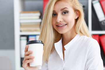 Smiling business woman drinking coffee from a paper cup in the office portrait looking into the camera holding in her hand and resting during a break