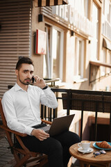 Handsome businessman talking on the phone and using a laptop for business online while sitting at the cafe table