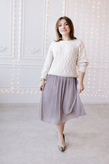 beautiful young woman in a white sweater, skirt and high heels on a background of white wall and christmas garlands.