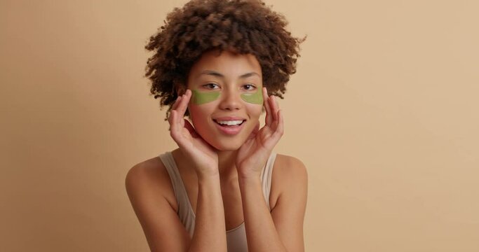 Beautiful glad African American woman applies eyes pads touches face gently shows her well groomed complexion and healthy skin poses against brown background. Spa face care cosmetology concept