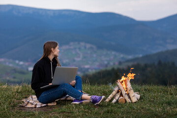 Fototapeta na wymiar young female freelancer working on laptop in the mountains in the evening. Tourist girl sitting near campfire and having fun. Copy space.