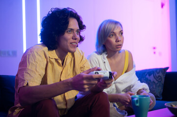 Young cheerful Caucasian husband and wife play videogames against each other at home enjoying leisure time