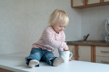 Two-year-old child in the kitchen with cup in his hands. Portrait of blonde toddler in the kitchen