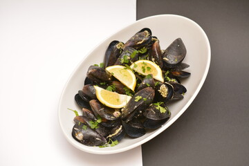 Black mussels on a white oval plate. Decorated with lemon wedges. Located on a black and white background. Can be used in the restaurant menu.
