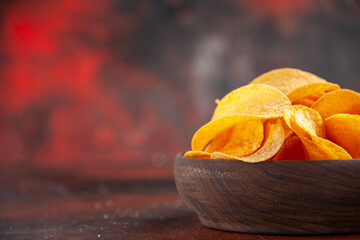 Side view of homemade delicious crispy potato chips in a small brown bowl on the left side of dark background