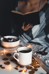 The woman is wrapped in a blanket and holds a book in her hands. cup of coffee . Winter comfort, garlands, Christmas mood. Stylish manicure