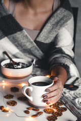 The woman is wrapped in a blanket and holds a cup of coffee in her hands. Winter comfort, garlands, Christmas mood. Stylish manicure