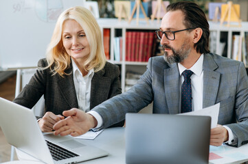 Mature business coworkers discussing about cooperation. Influential senior man consults beautiful woman on working process. Confident colleagues are sitting at the work desk in office, using laptop