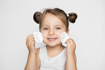 Fototapeta premium Studio shot of pretty kid girl, cleansing her face by cotton pads, posing at camera with happy smile, enjoying skincare procedures. Close up portrait on isolated white background
