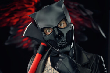 a man in a demon skull mask in a leather cloak with an axe on a background of black and red wings