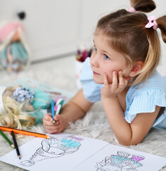 Cute pretty small girl in home clothing dreaming and coloring doll drawing with colorful pencils at home. Happy childhood, cheerful lifestyle, games, comfortable pastime, hobby concept