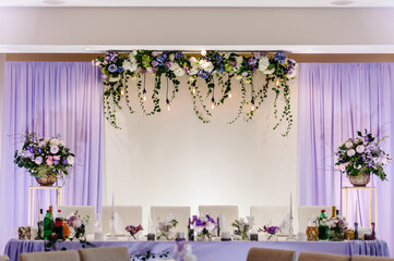 Festive table, arch, stands decorated with composition of violet, purple, pink flowers and greenery, candles in the banquet hall. Table newlyweds in the banquet area on wedding party.