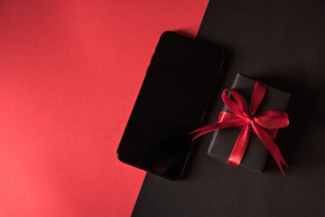 Black Friday sale shopping concept, Top view of gift box wrapped in black paper and black bow ribbon and modern mobile smartphone, studio shot on red and dark background