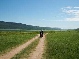 Young man rides a motorbike carrying equipment for a long vacation. In green meadows by lakes and valleys
