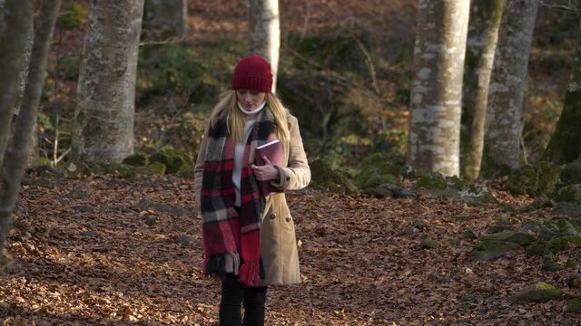 Young Woman in Red Hat and Scarf Holds Book Walking in Autumn Forest