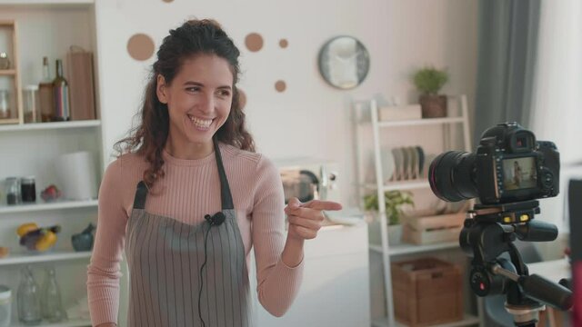 Waist up shot of young Caucasian female influencer wearing apron standing in kitchen, talking, smiling, pointing with finger gun hand sign on camera set up in front of her