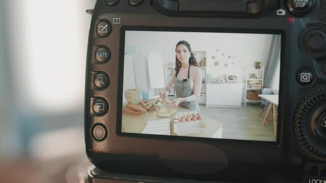 Screen of semi-professional camera with running video of young Caucasian woman standing by kitchen table cooking and talking