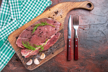 Top view of red meat on wooden cutting board on green stripped towel and garlic fork and knife on dark background