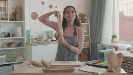 Medium POV of cheerful Caucasian young woman dressed in apron standing by table with utensils in kitchen, cooking and stretching dough, smiling, speaking, looking on camera