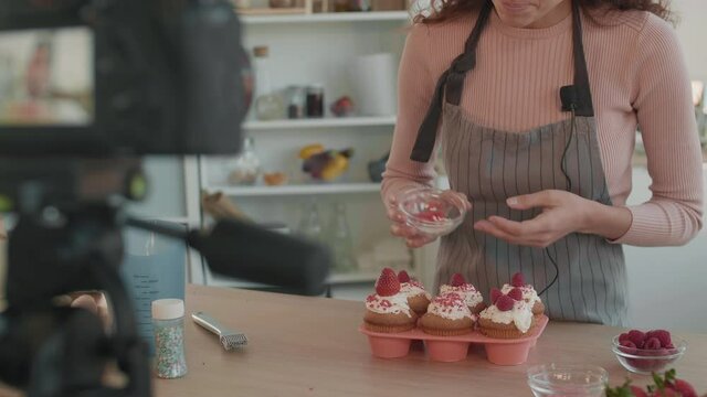 Cropped person in apron standing at table in kitchen, decorating muffins with fresh berries and sprinkles, blurred camera in front filming process