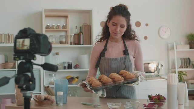 Medium shot of young Caucasian woman in apron coming to table holding baking sheet with freshly baked muffins, smelling them and showing on camera, set on tripod in front