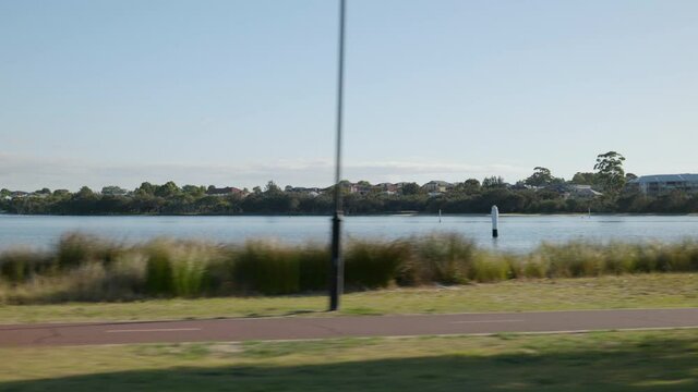 Move along peaceful Canning River with houses, trees in background. Bike path and trees in foreground. Rowers resting on water