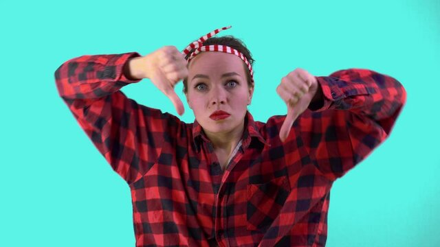 Pretty woman dressed in pin-up style, dressed in a red shirt in a cage and a bandage on her head showing thumbs down gesture on a blue background in the studio.
