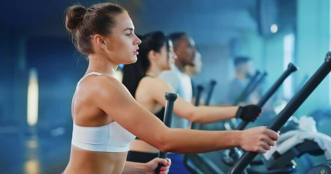 Attractive female athlete fitness coach training on cross trainer exercise bike with team sports smiling positively at camera in modern gym interior.