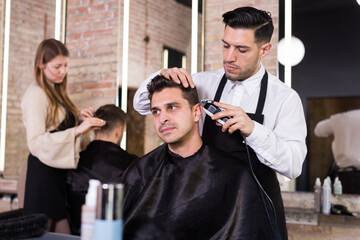 Professional hairdresser man doing styling of handsome guy with electric hair clipper at barber shop
