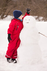 Fototapeta na wymiar Portrait of a cute boy in red winter jumpsuit making a big snowman in the park. Child having fun outdoors. The winter vacation. Early childhood development concept.