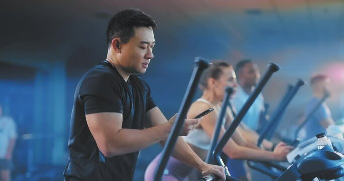 Asian cheerful man trainingin sports club on eliptical bike using smartphone recording video messages for girlfriend rejoicing. Sportsman. Busy lifestyle. Nomophobia.