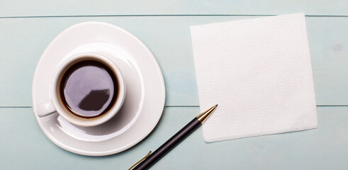 Obraz na płótnie Canvas On a light blue wooden background, a white cup of coffee, an empty napkin for notes and a pen. Top view with copy space