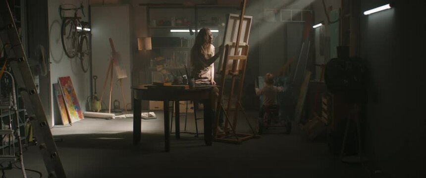 WIDE Female artist working in her garage studio, little daughter playing around . Shot with 2x anamorphic lens