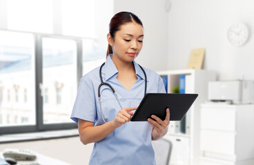 medicine, profession and healthcare concept - asian female doctor or nurse in blue uniform with tablet pc computer and stethoscope over hospital background