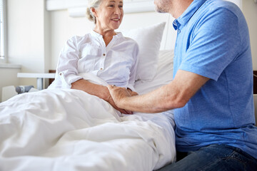 Obraz premium medicine, healthcare and people concept - happy senior man visiting and cheering his woman lying in bed at hospital ward