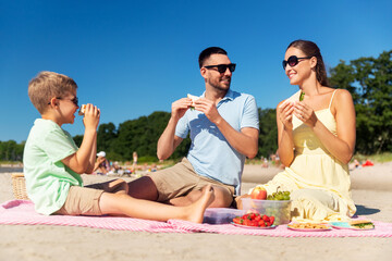 family, leisure and people concept - happy mother, father and little son having picnic on summer beach and eating sandwiches