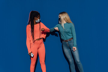Two women of different ethnicity wearing masks greeting each other with their elbows in blue background