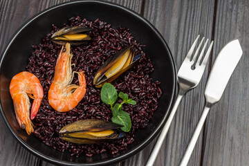 Shrimp and mussels with black rice in bowl. Fork and knife on table