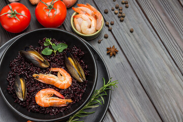 Shrimps and mussels with black rice on plate. Tomatoes, shrimps in bowl. Dark wooden background
