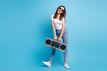 Full size photo of young happy cheerful positive smiling girl in sunglasses hold boombox isolated on blue color background