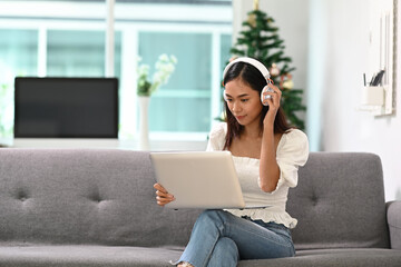 Naklejka premium A young woman wearing headphone enjoy watching media on laptop while sitting on a sofa in the living room.